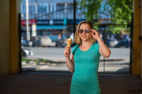 Eine glückliche Frau in einem türkisfarbenen Kleid steht im Hof und isst an einem warmen Sommertag eine Waffel. Die schöne Blondine mit Sonnenbrille genießt Eis beim Gehen. Sommer-Dessert. — Stockfoto