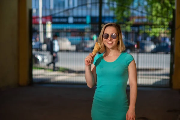 Eine glückliche Frau in einem türkisfarbenen Kleid steht im Hof und isst an einem warmen Sommertag eine Waffel. Die schöne Blondine mit Sonnenbrille genießt Eis beim Gehen. Sommer-Dessert. — Stockfoto