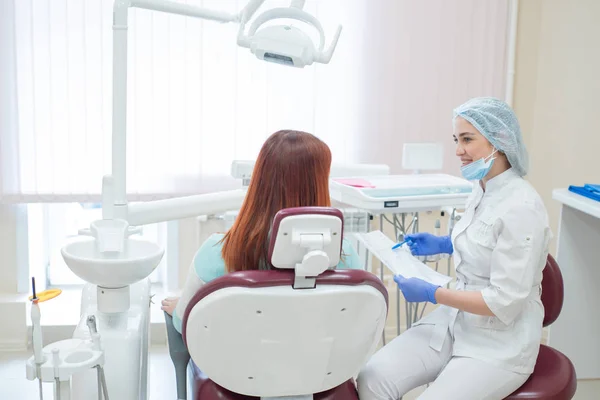 Giovane bella dentista che parla con una paziente in studio. Una donna dai capelli rossi siede su una sedia con un ortodontista e ascolta gli appuntamenti. Servizi dentistici. Vista dal retro — Foto Stock