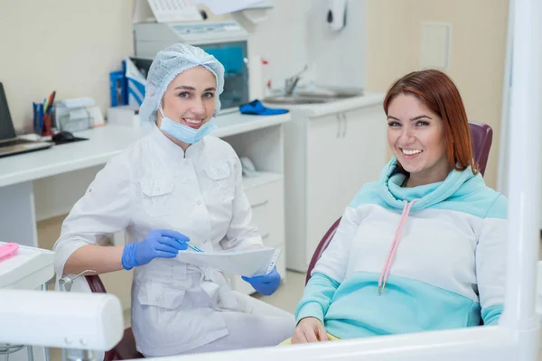 Giovane bella dentista femminile e paziente felice stanno guardando direttamente nella fotocamera. Donna dai capelli rossi siede su una sedia con un ortodontista e sorride. Servizi dentistici . — Foto Stock