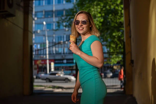 Eine glückliche Frau in einem türkisfarbenen Kleid steht in einem gewölbten Gang und isst an einem warmen Sommertag einen Waffelkegel. Die schöne Blondine mit Sonnenbrille genießt Eis beim Gehen. Sommer-Dessert. — Stockfoto