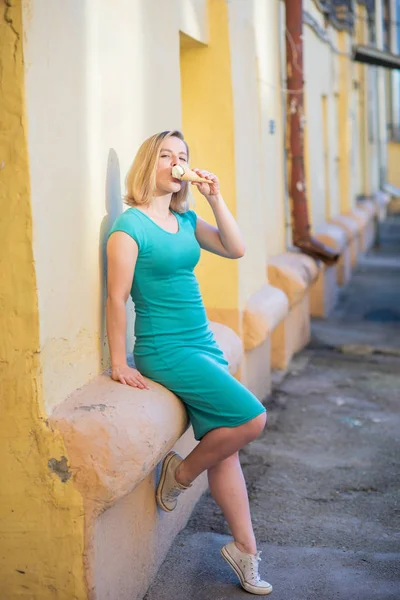 Портрет милой девушки в зеленом платье гуляет по двору и ест мороженое. Красивая блондинка, прислонившись к стене дома, ест десерт в прекрасный летний день. Жизнь - это приятный отдых . — стоковое фото