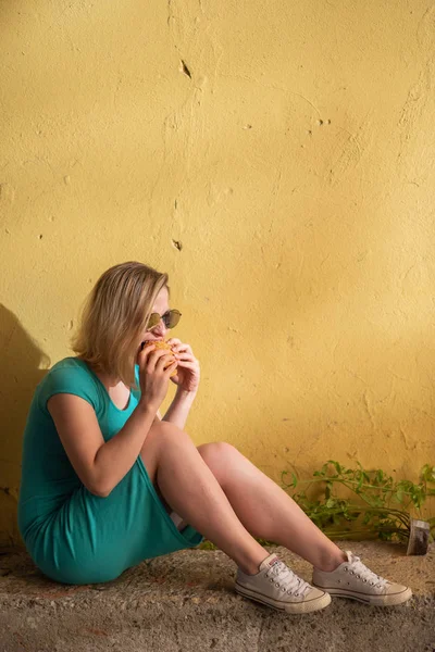 Uma mulher feliz em um vestido turquesa senta-se na calçada e come um hambúrguer em um dia quente de verão. Loira bonita em óculos de sol goza de junk food na parede amarela de um edifício residencial . — Fotografia de Stock