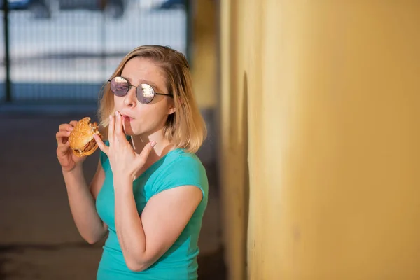 Portrét roztomilé dívky v zelených šatech venku a jíst Burger v teplém letním dnu. Krásná blondýnka v slunečních brýlích má na žluté stěně na ulici jídlo na smetí. Špatné stravní návyky. — Stock fotografie