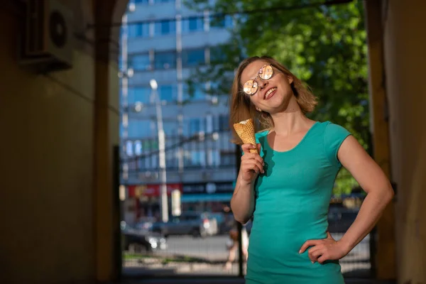 Eine glückliche Frau in einem türkisfarbenen Kleid steht in einem gewölbten Gang und isst an einem warmen Sommertag einen Waffelkegel. Die schöne Blondine mit Sonnenbrille genießt Eis beim Gehen. Sommer-Dessert. — Stockfoto