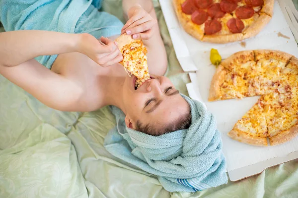 Linda chica con una toalla en la cabeza come pizza en la cama. Mujer joven comiendo pizza en la cama. La vida es un placer, un cuerpo positivo. Amor por la comida italiana. Hábitos alimenticios, adicción a la comida rápida . — Foto de Stock