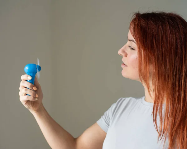 Ritratto di una donna dai capelli rossi con un ventilatore tascabile in mano. Profilo di una ragazza rinfrescata nel calore da un vento soffiato da un dispositivo elettrico wireless. Godetevi il flusso d'aria fresca dal condizionatore . — Foto Stock