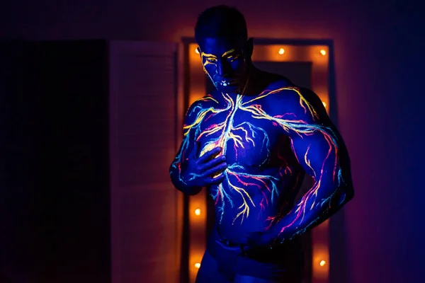 Modelli UV body art del sistema circolatorio sul corpo di un uomo. Sul busto di un atleta muscolare, vene e arterie sono disegnate con coloranti fluorescenti. Bodybuilder in piedi accanto allo specchio con lampade . — Foto Stock