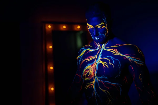 Modelli UV body art del sistema circolatorio su un corpo umano. Sul busto di un atleta muscolare, vene e arterie sono disegnate con coloranti fluorescenti. Bodybuilder in piedi accanto allo specchio con lampade. — Foto Stock