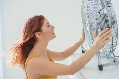 Hardal tulumlu güzel kızıl saçlı bir kadının portresi büyük bir elektrik fanından gelen soğutma esintisinin tadını çıkarıyor. Kız yaz aylarında bunaltıcı sıcağında yenilenir. Saç rüzgardan gelişir.