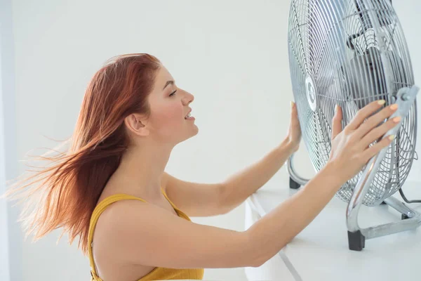 Retrato de una hermosa mujer pelirroja con monos de mostaza disfrutando de la brisa refrescante de un gran ventilador eléctrico. La chica se refresca en el calor sofocante verano. El cabello se desarrolla por el viento . — Foto de Stock