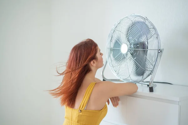 Büyük bir elektrikli fan soğutma esinti zevk bir hardal tulum güzel bir kırmızı saçlı kadının portresi. Gülümseyen bir kız sıcak yaz sıcağında tazeler. Saç rüzgar gelişir — Stok fotoğraf