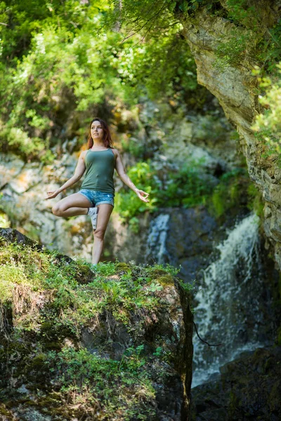 Rødhåret kvinne i shorts som gjør yoga ved fossen. En jente står i et tre i skogen. – stockfoto