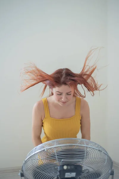 Portrét nádherné rudovlasé ženy v hořčičné kombinéze, která si vychutnávala chladicí vánek od velkého elektrického vějíře. Usměvavá dívka se v horkém letním žáru osvěžuje. Vlasy se vyvíjejí od větru — Stock fotografie
