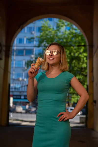 Счастливая женщина в бирюзовом платье стоит в арочном проходе и ест вафельный конус в теплый летний день. Красивая блондинка в солнечных очках наслаждается мороженым во время ходьбы. Летний десерт. — стоковое фото