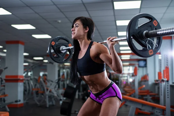 Ein kräftiges, muskulöses dunkelhaariges Mädchen macht Kniebeugen mit einer hellen Langhantel. Fitnessstudio — Stockfoto