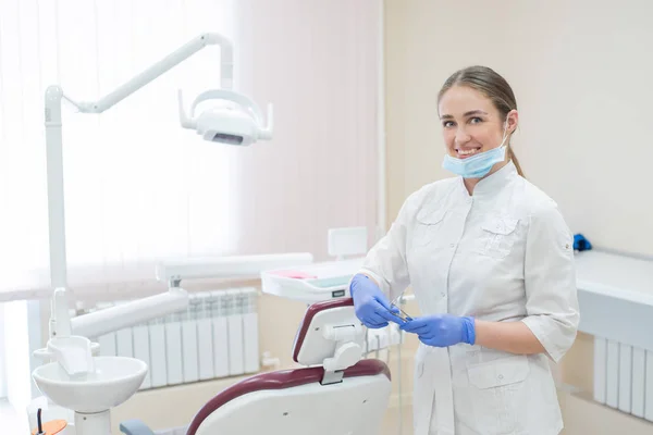 Attraente sorridente medico donna in uniforme bianca sul posto di lavoro. Giovane bella dentista donna in maschera e guanti è in piedi nello studio vicino alla poltrona con gli strumenti in mano — Foto Stock