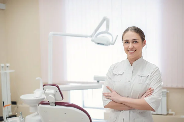 Atrakcyjny uśmiechnięty żeński lekarz w białym mundurze w miejscu pracy. Młoda piękna kobieta dentysta brunetka w biurze przez krzesło — Zdjęcie stockowe