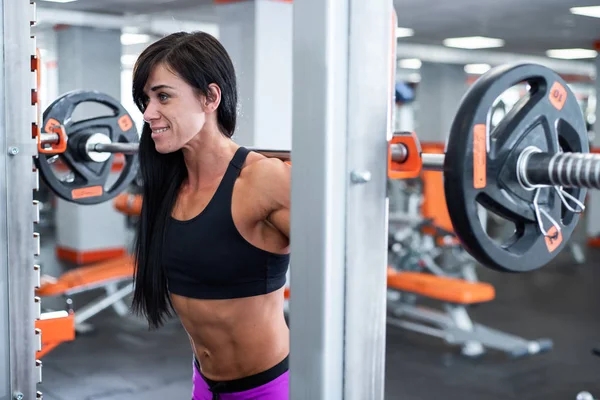 Ein kräftiges, muskulöses dunkelhaariges Mädchen macht Kniebeugen mit einer hellen Langhantel. Fitnessstudio — Stockfoto