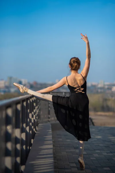 Μπαλαρίνα σε μια φούστα που ποζάρει κοντά στον φράχτη. Όμορφη νεαρή γυναίκα με μαύρο φόρεμα και το χορό που χορεύουν έξω. Πανέμορφη μπαλαρίνα δείχνει εκπληκτική τέντωμα. — Φωτογραφία Αρχείου