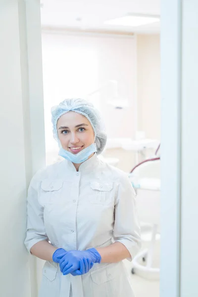 Atractiva doctora sonriente en uniforme blanco con guantes está de pie en la puerta de la oficina. dentista en el pasillo de una clínica con gorra y máscara . —  Fotos de Stock