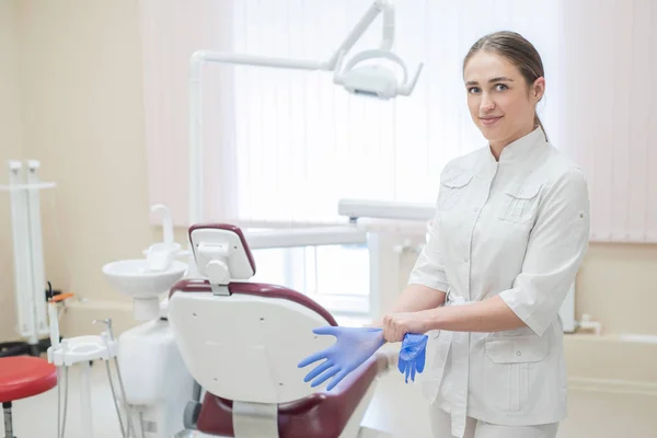 La atractiva doctora en uniforme blanco se pone guantes. Joven hermosa mujer dentista morena en la oficina por la silla con guantes . —  Fotos de Stock