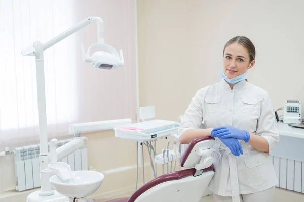 Attraente sorridente medico donna in uniforme bianca sul posto di lavoro. Una giovane bella dentista in maschera e guanti è in piedi nello studio vicino alla sedia — Foto Stock