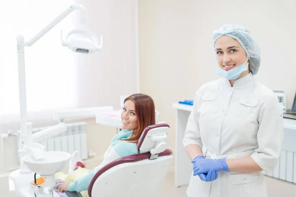 Joven hermosa dentista y paciente feliz están mirando directamente a la cámara. Mujer pelirroja se sienta en una silla con un ortodoncista y sonríe. Servicios dentales . — Foto de Stock