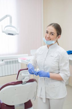 İşyerinde beyaz üniformalı çekici gülümseyen kadın doktor. Maskeli ve eldivenli genç güzel kadın diş hekimi elinde aletlerle koltuğun yanında ofiste duruyor.