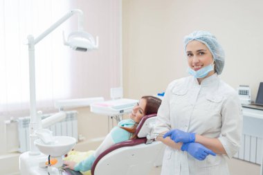 Genç güzel kadın dişçi geri ofiste hastaların arkasında duruyor. Kızıl saçlı kadın bir ortodontist ile bir sandalyede oturur ve gülümsüyor. Diş Hekimliği Hizmetleri. Arkadan görünüm