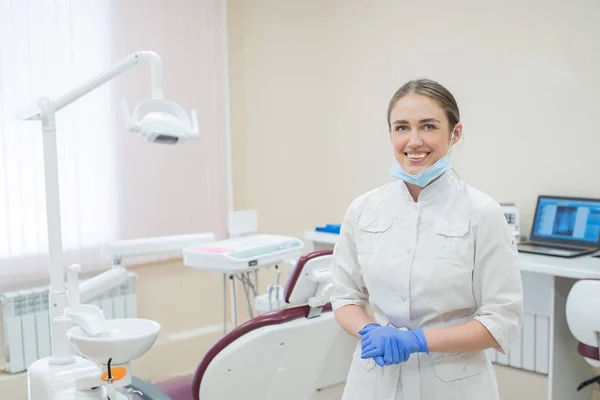 Attraente sorridente medico donna in uniforme bianca sul posto di lavoro. Una giovane bella dentista in maschera e guanti è in piedi nello studio vicino alla sedia — Foto Stock