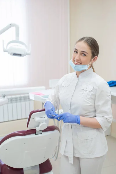 Attraente sorridente medico donna in uniforme bianca sul posto di lavoro. Giovane bella dentista donna in maschera e guanti è in piedi nello studio vicino alla poltrona con gli strumenti in mano — Foto Stock