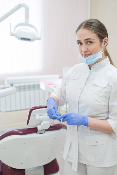 有吸引力的微笑女医生在工作服的白色制服。年轻美丽的女牙医戴着面具和手套站在办公室附近的扶手椅上，手里拿着工具 — 图库照片
