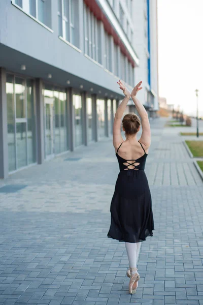 Μπαλαρίνα σε μια φούστα που ποζάρει στο φόντο ενός κτηρίου κατοικιών. Όμορφη νεαρή γυναίκα με μαύρο φόρεμα και Pointe παπούτσια χορεύοντας μπαλέτο έξω. — Φωτογραφία Αρχείου