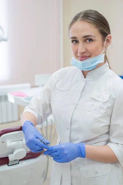 Atractiva doctora sonriente con un uniforme blanco en el lugar de trabajo. Joven hermosa dentista con máscara y guantes está de pie en la oficina cerca del sillón con herramientas en las manos — Foto de Stock