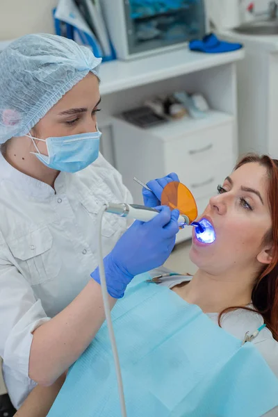 Dentista femenina fija y seca los rellenos dentales con luz. Hermosa dentista femenina poniendo un relleno en un diente con caries de una paciente de cabello rojo —  Fotos de Stock