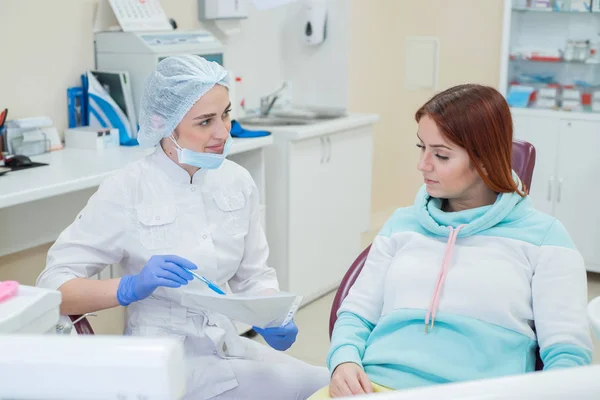 Joven dentista hermosa hablando con una paciente en el consultorio. Una mujer pelirroja se sienta en una silla con un ortodoncista y escucha las citas. Servicios dentales —  Fotos de Stock