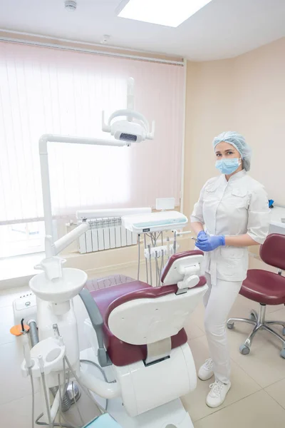 Dentysta dziewczyna, w białych ubrań, maska i czapka stoi obok nowoczesnego fotela w dużym, prostym, gabinecie lekarskiej — Zdjęcie stockowe