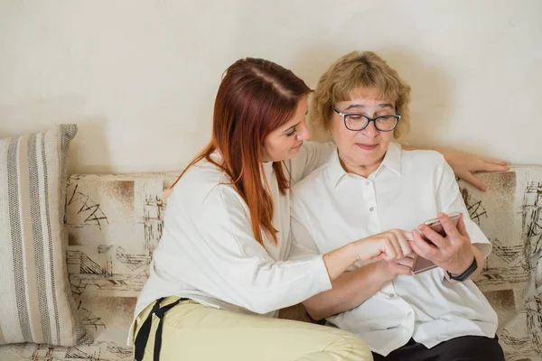 Kızıl saçlı kadın yaşlı bir anneye akıllı telefonu kullanmayı öğretir. Anne ve yetişkin kızı beyaz bluz giymiş oturma odasında kanepede oturuyor ve telefonda fotoğrafları izliyor. — Stok fotoğraf