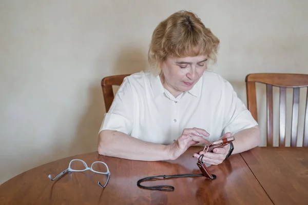 테이블에 앉아 흰색 블라우스에 아름 다운 노인 여자와 전화에 메시지를 씁니다. 은퇴 한 여성은 점수를 득점하고 스마트 폰에서 메시지를 읽습니다. — 스톡 사진