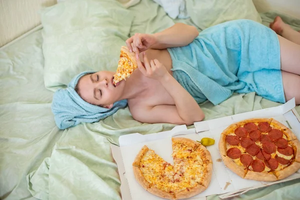 Schattig meisje met een handdoek op haar hoofd eet pizza in bed. Jonge vrouw eten pizza in bed. Het leven is een genot, lichaam positief. Liefde voor Italiaans eten. Voedingsgewoonten, verslaving aan Fast Food. — Stockfoto