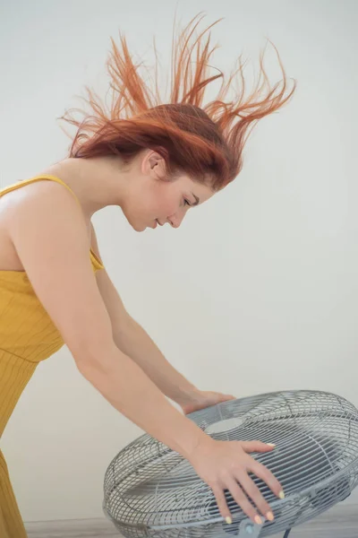 Portret pięknej rudowłosy Kobieta w kombinezon musztardowy korzystających z chłodzenia bryza z dużego wentylatora elektrycznego. Uśmiechnięta dziewczyna odświeży się w gorącym letnim upale. Włosy rozwijają się od wiatru — Zdjęcie stockowe