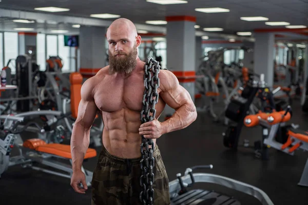 Een kale, bebaarde bodybuilder met grote reliëf spieren heeft een zware dikke ketting in zijn handen. Lichte fitnessruimte. — Stockfoto