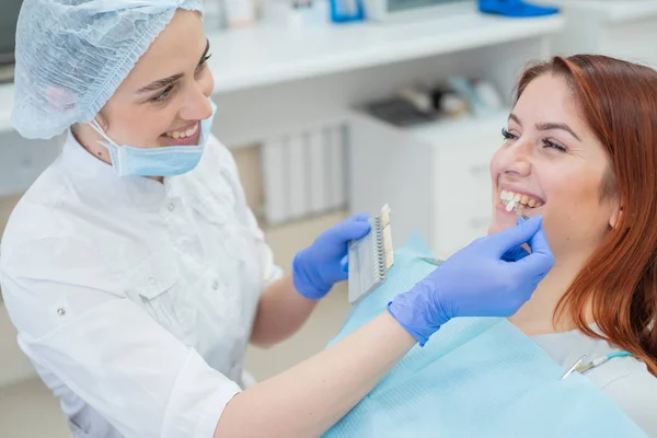 Controllare e selezionare il colore dei denti sulla sedia del dentista. Il dentista fa il processo di trattamento di una bella giovane ragazza dai capelli rossi. Selezione di un impianto dentale . — Foto Stock