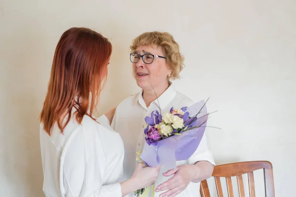 Daughter giving flowers to adult mother. Daughter gives flowers to her mother and hugs her. Spending time together, celebrating at home on weekends. Mothers Day