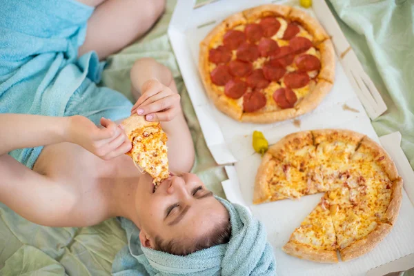 Linda chica con una toalla en la cabeza come pizza en la cama. Mujer joven comiendo pizza en la cama. La vida es un placer, un cuerpo positivo. Amor por la comida italiana. Hábitos alimenticios, adicción a la comida rápida . — Foto de Stock