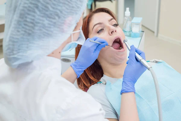 Una atractiva doctora sonriente en uniforme blanco examina a una paciente de cabello rojo. Dentista produce trata los dientes con un taladro . —  Fotos de Stock