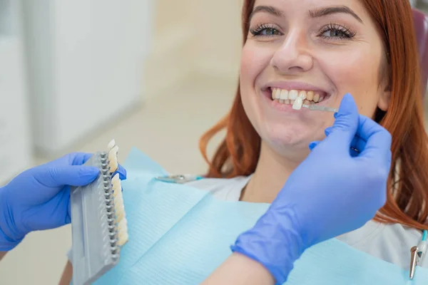 Controllare e selezionare il colore dei denti sulla sedia del dentista. Il dentista fa il processo di trattamento di una bella giovane ragazza dai capelli rossi. Selezione di un impianto dentale. Primo piano . — Foto Stock