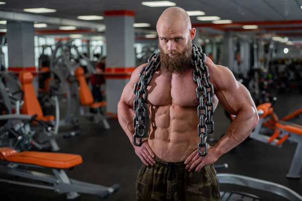 Un culturista calvo y barbudo con grandes músculos en relieve sostiene una gruesa cadena en sus manos. Gimnasio brillante . — Foto de Stock