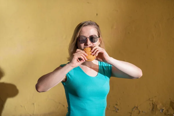 Портрет милой девушки в зеленом платье, идущей на улицу и поедающей бургер в теплый летний день. Красивая блондинка в солнечных очках наслаждается нездоровой пищей на желтой стене на улице. Неправильные привычки питания . — стоковое фото